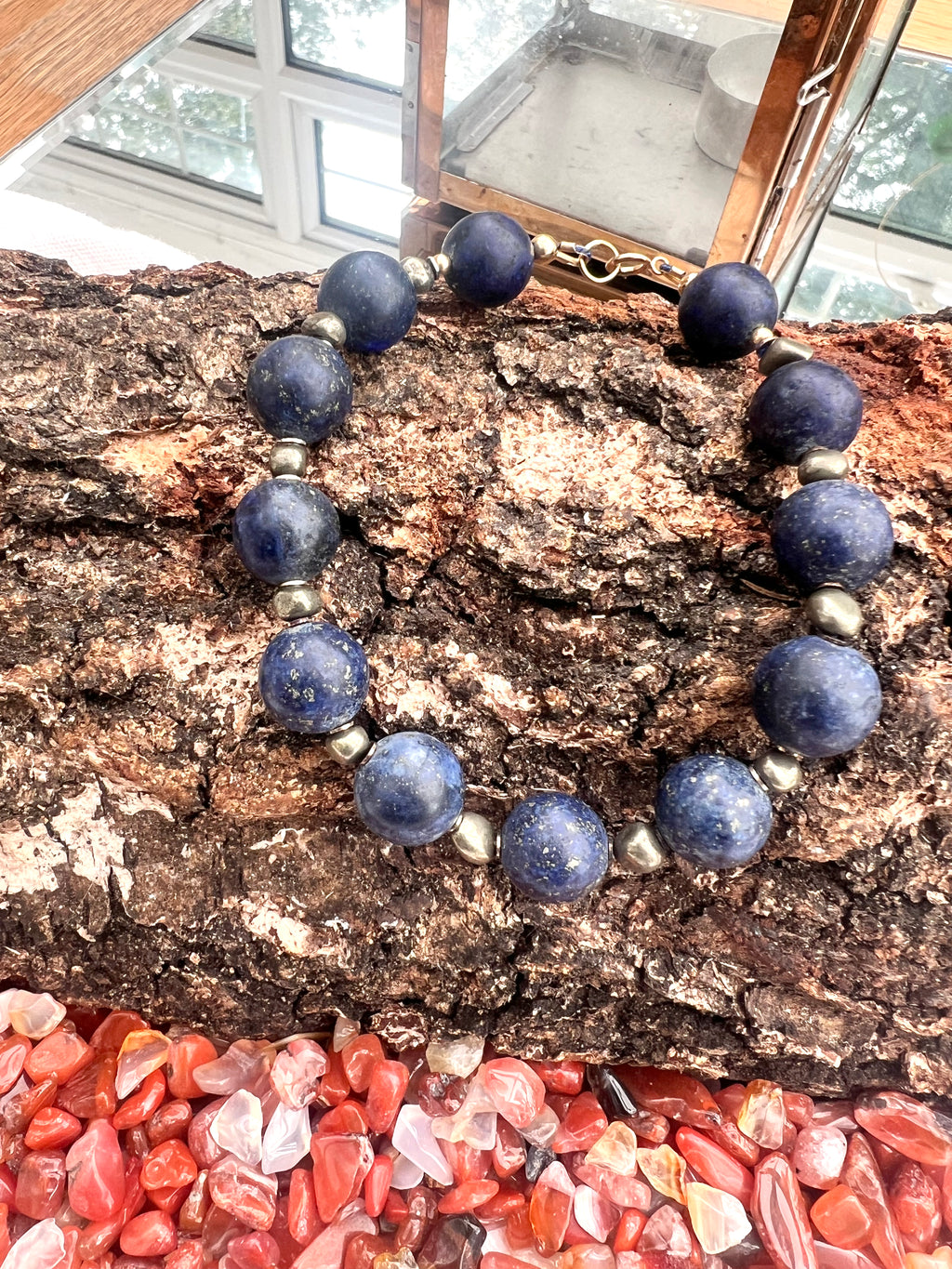 A chunky bracelet made of dark blue lapis lazuli stones with small irregular pyrite, displayed on a piece of textured wood above a pile of carnelian gemstones. A small paper tag reads alaraJade