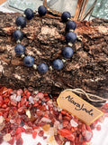 A chunky bracelet made of dark blue lapis lazuli stones with small irregular pyrite, displayed on a piece of textured wood above a pile of carnelian gemstones. A small paper tag reads alaraJade
