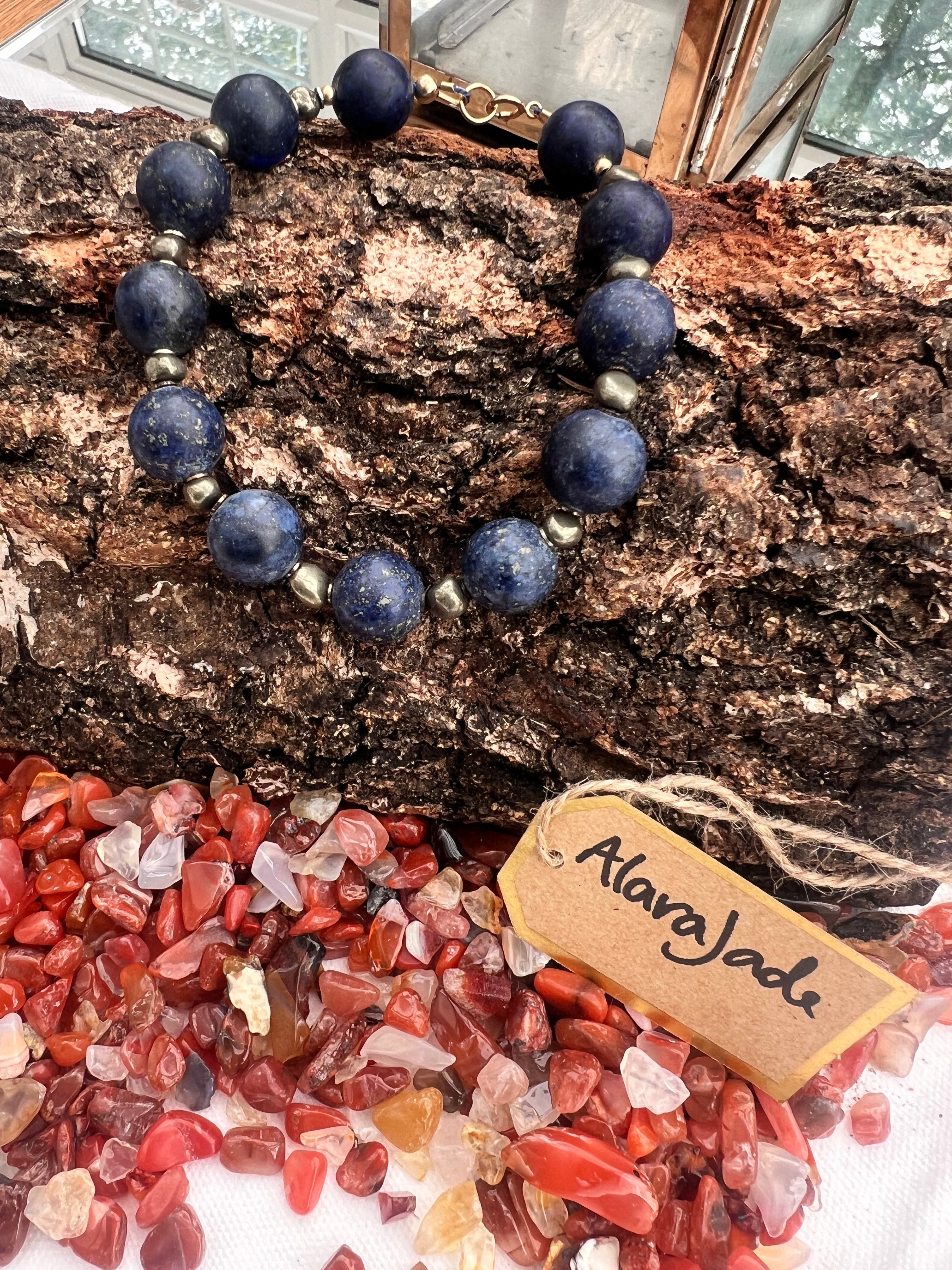 A chunky bracelet made of dark blue lapis lazuli stones with small irregular pyrite, displayed on a piece of textured wood above a pile of carnelian gemstones. A small paper tag reads alaraJade