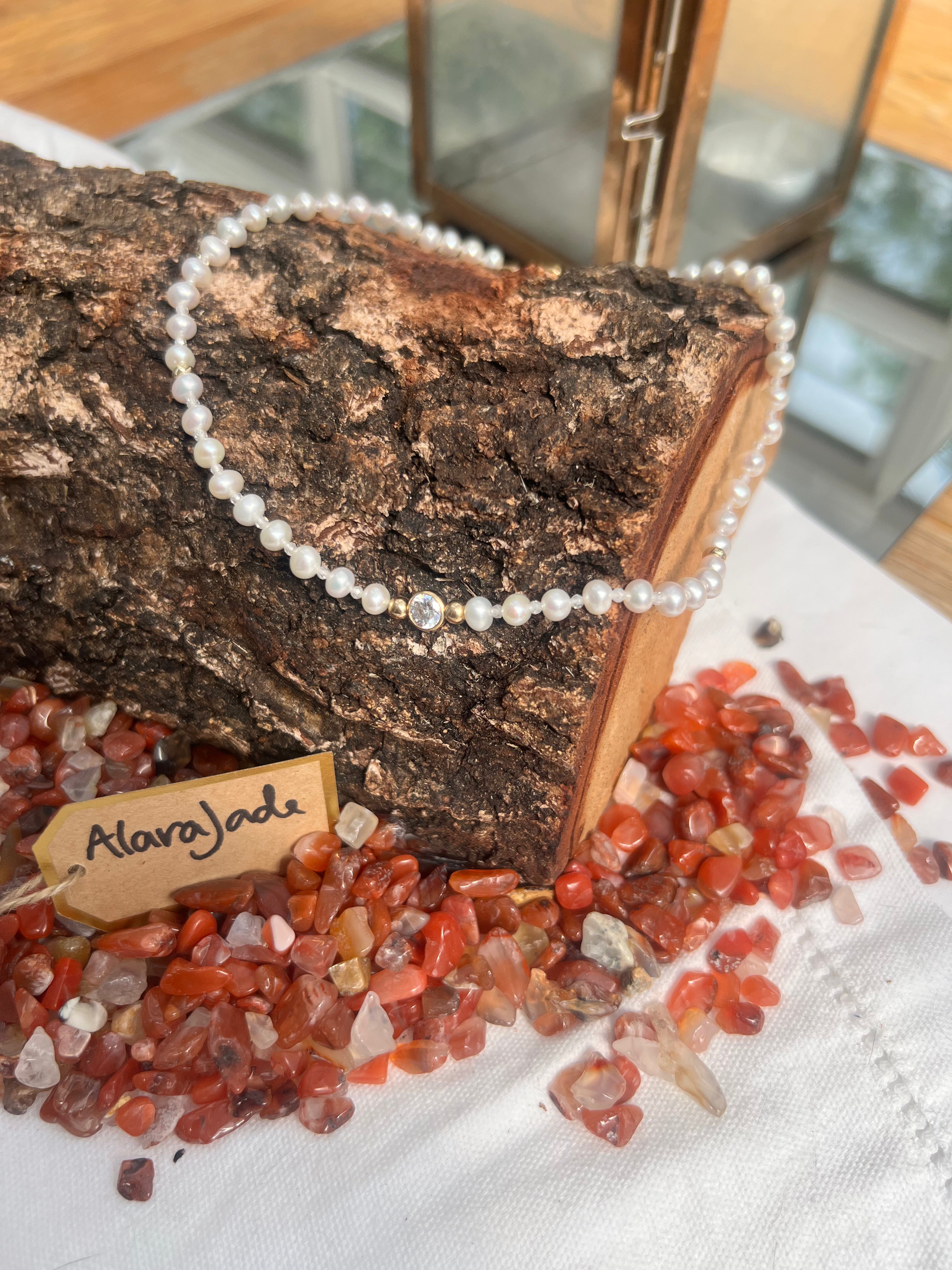A white pearl necklace with a central clear stone and a small gold accent bead, draped over a piece of rough wood. The necklace is displayed on a white cloth surrounded by red and orange carnelian chips, with a small wooden tag reading 'Alarajade'.