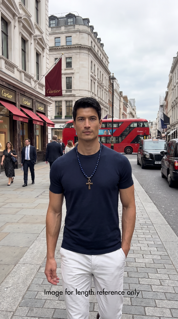 A man wearing a navy blue crewneck t-shirt and white pants stands on a city street in London wearing a necklace with dark blue lapis lazuli round beads, small pyrite spacer beads, and a golden-toned ankh pendant.