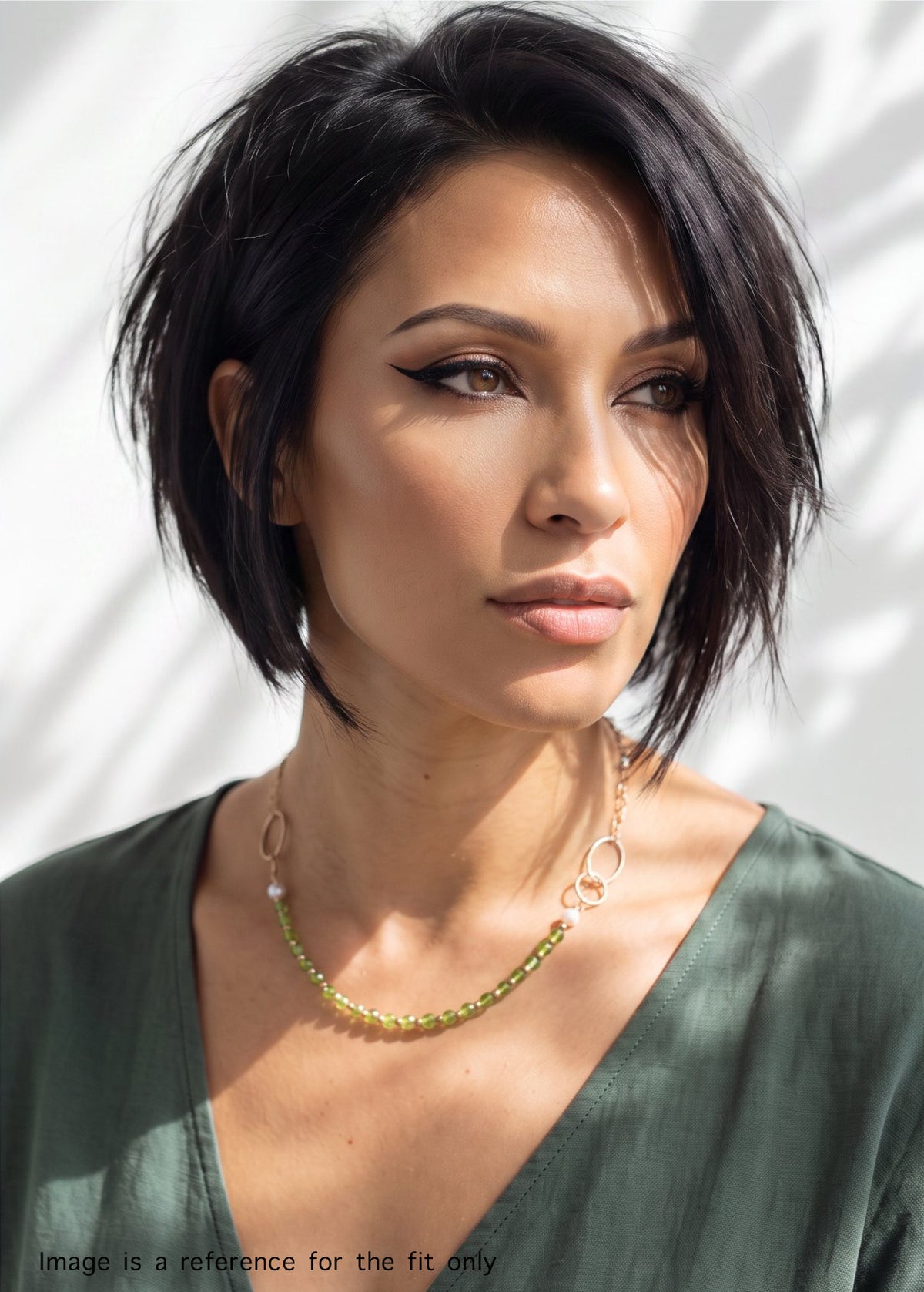 A woman with short dark hair wearing a delicate peridot and gold chain necklace, paired with a green top against a white background with soft leaf shadows.