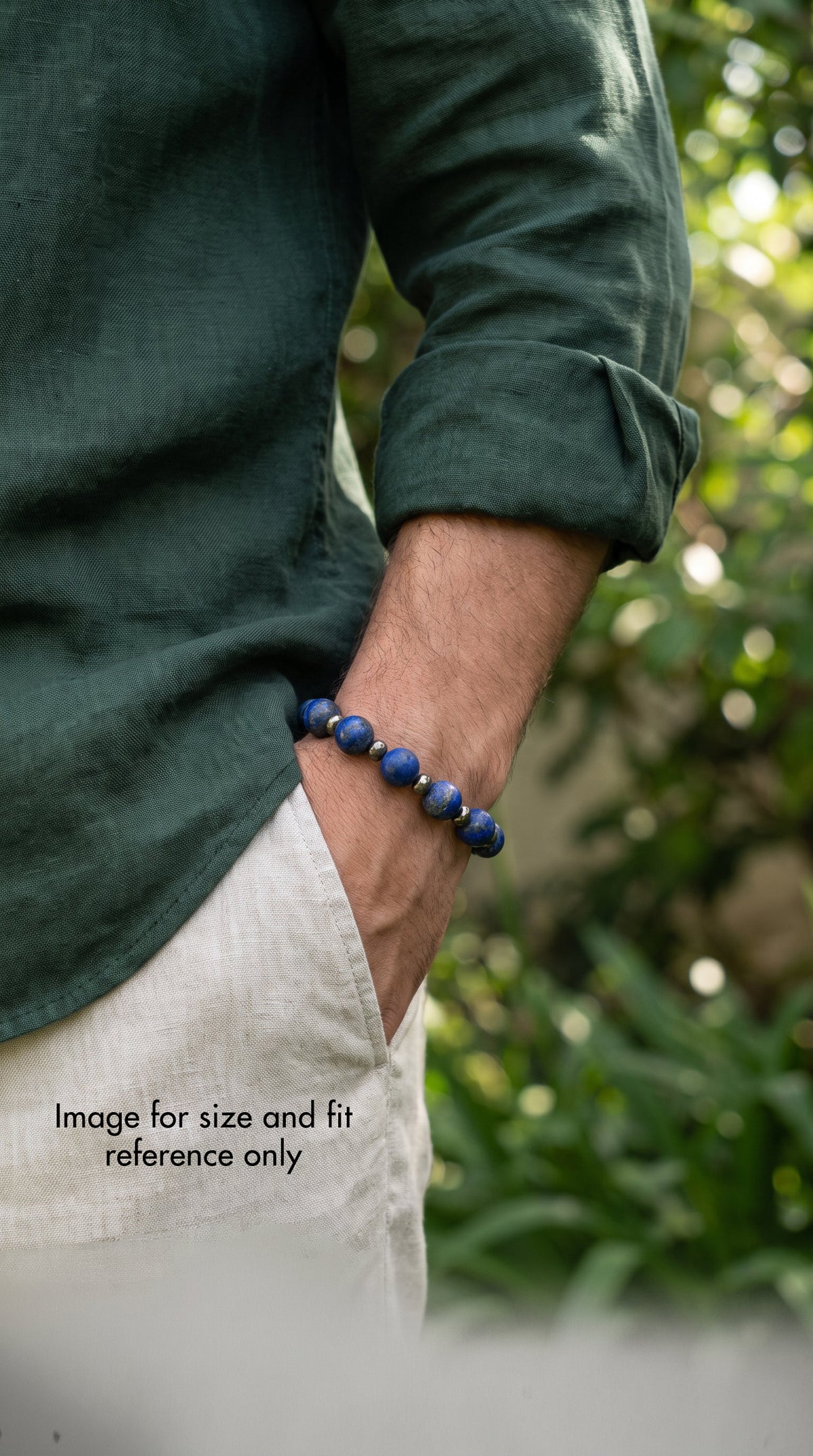 A close-up lifestyle shot of a man wearing a dark green linen shirt and off-white trousers, with his hand in his pocket. He is wearing a dark blue lapis lazuli and pyrite bracelet on his wrist.