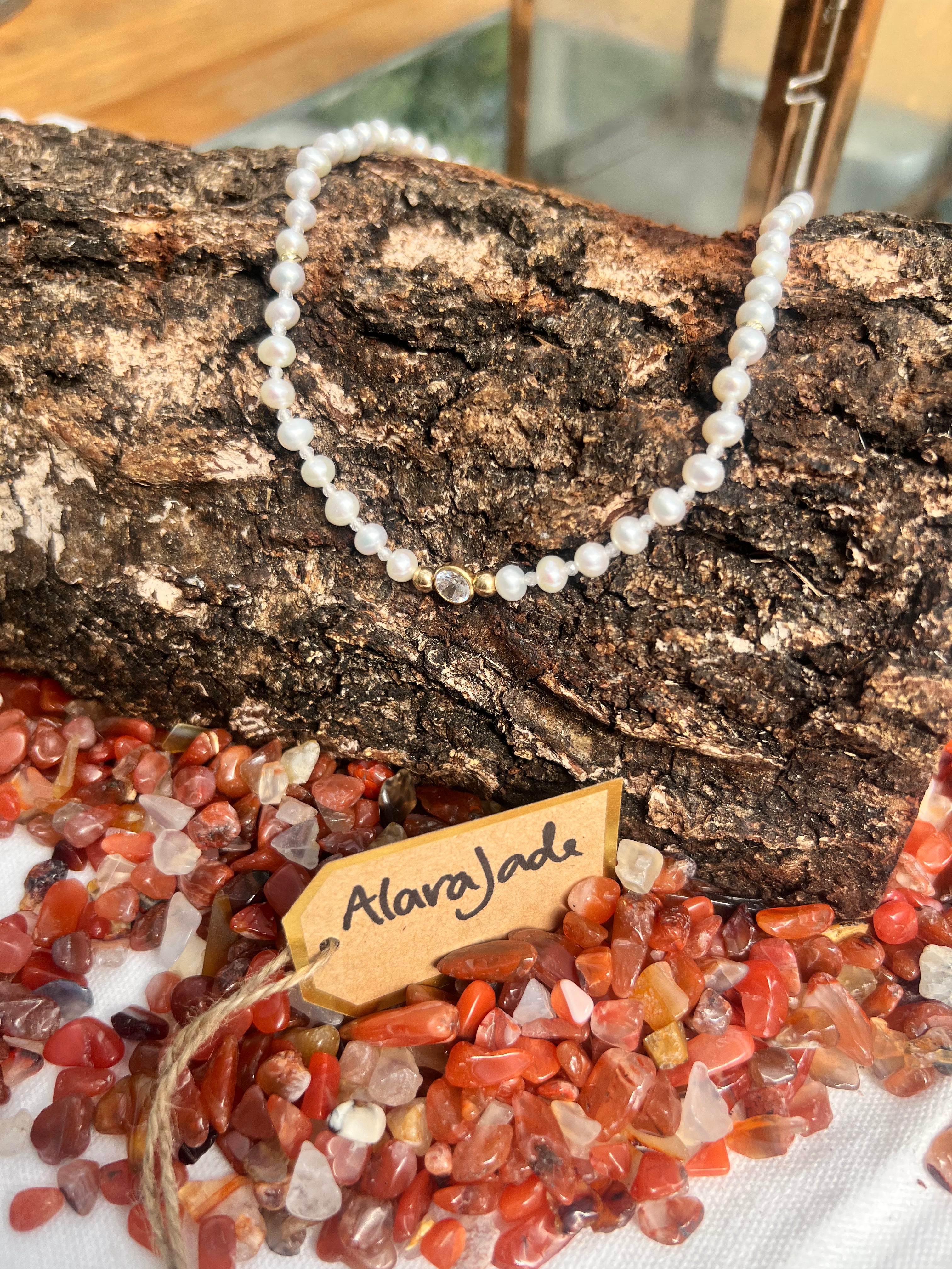 A white pearl necklace with a central clear crystal centrepiece, draped over a piece of textured tree bark. The necklace is displayed on a white cloth surrounded by red and orange polished gemstones, with a small wooden tag reading 'AlaraJada' in script.