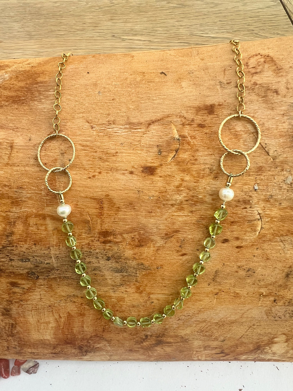 A gold-filled chain necklace with two interlocking rings on each side, a single pearl bead on each side, and a strand of faceted light green coin peridot, displayed on a rustic wooden surface.