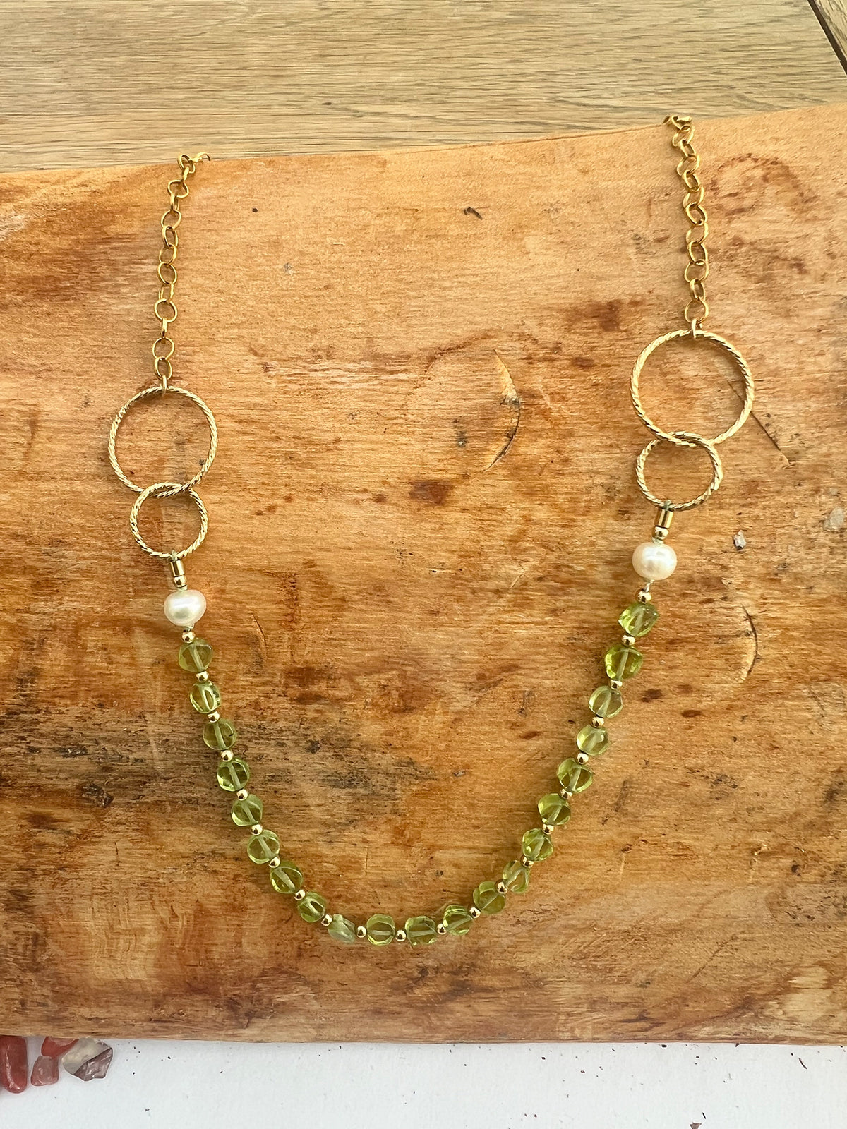 A gold-filled chain necklace with two interlocking rings on each side, a single pearl bead on each side, and a strand of faceted light green coin peridot, displayed on a rustic wooden surface.