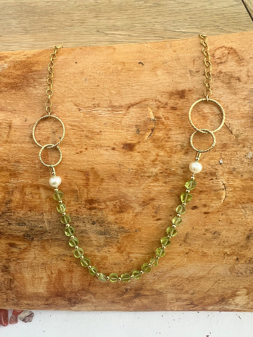 A gold-filled chain necklace with two interlocking rings on each side, a single pearl bead on each side, and a strand of faceted light green coin peridot, displayed on a rustic wooden surface.